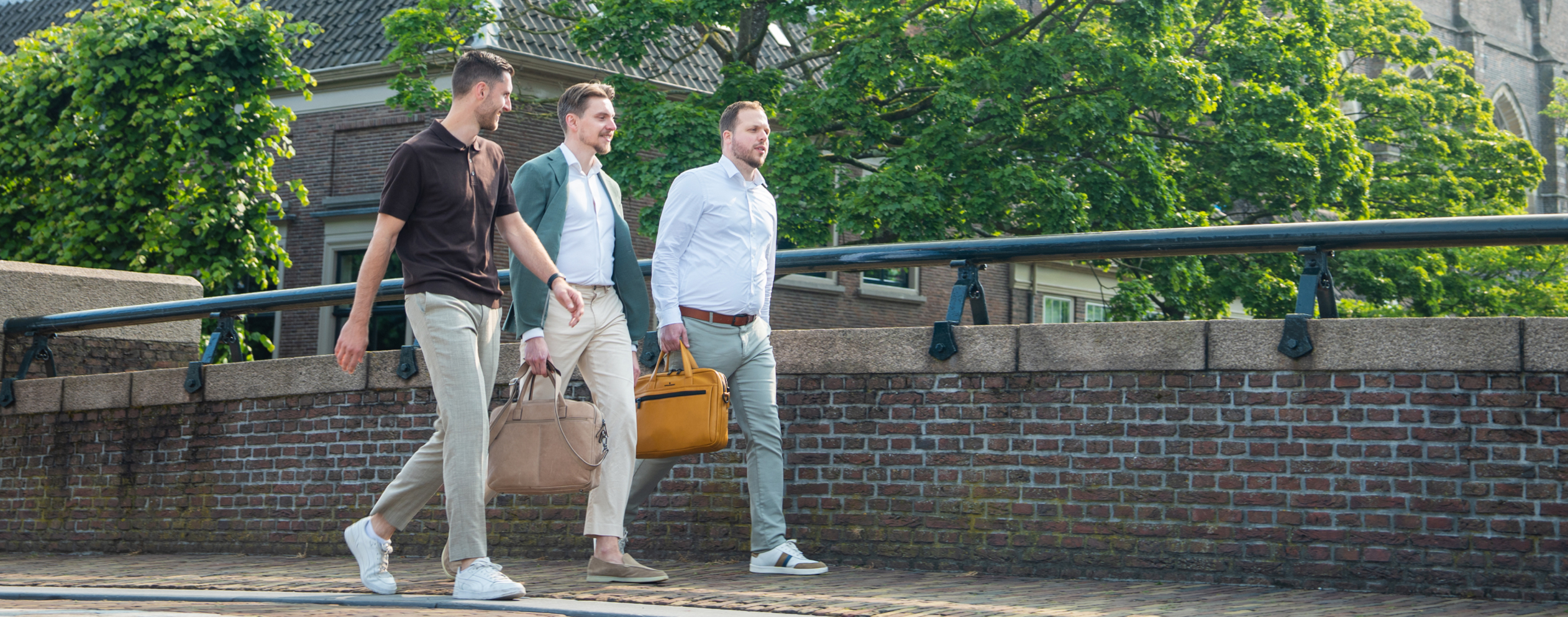 Three men casually strolling across a bridge, surrounded by greenery and a clear blue sky.
